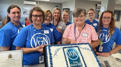 In an office setting, VON staff (women) gather around to pose for a photo with a large cake that has the VON logo with tag that says Live Every Day. on it. 